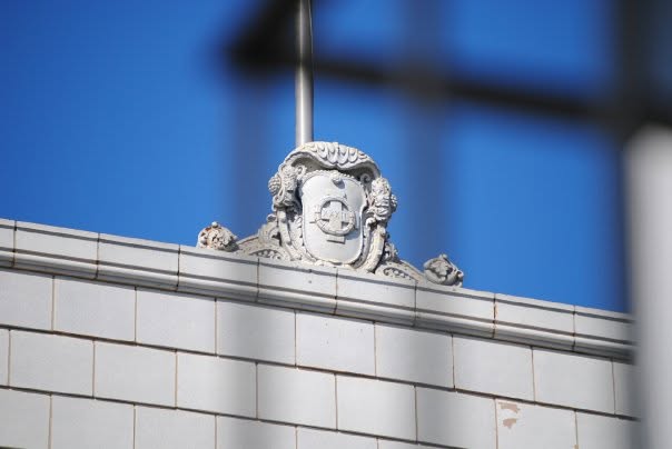 An ornate stone architectural crest above a tiled building facade, framed against blue reflected glass